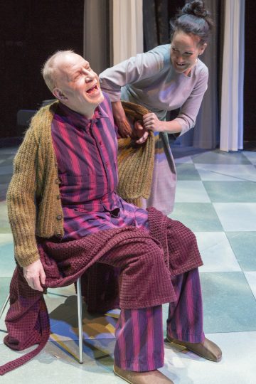 Raidalliseen pyjamaan ja ruskeaan neuletakkiin pukeutunut vanhempi mies istuu tuolilla nauraen, kun hymyilevä nainen auttaa häntä pukemaan villapaidan päälleen. He ovat näyttämöllä, jossa on pehmeä valaistus ja ruudullinen lattia.