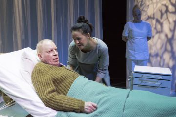 Iäkäs mies makaa sairaalasängyssä vihreällä peitteellä peitettynä. Huolestunut nainen kumartuu häntä kohti ja puhuu keskittyneesti, kun valkoisiin pukeutunut hoitaja seisoo taustalla ja tarkkailee tilannetta.