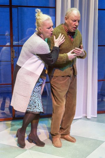 Vaaleatukkainen nainen auttaa tukemaan vanhempaa miestä, jolla on vihreä villapaita ja ruskeat housut. He seisovat sisätiloissa lasiovien ja valkoisten verhojen lähellä, ja molemmat vaikuttavat huolestuneilta ja tunteellisilta.