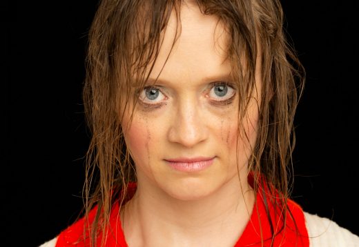 Nainen, jolla on märkä tukka ja tahriintunut ripsiväri, tuijottaa suoraan kameraan intensiivinen ilme kasvoillaan, päällään punavalkoinen toppi mustaa taustaa vasten.