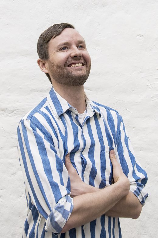 Lyhyet ruskeat hiukset ja partainen mies hymyilee seisoessaan valkoista seinää vasten. Hänellä on yllään sinivalkoinen pystyraidallinen paita ja kädet ristissä rintakehän päällä.
