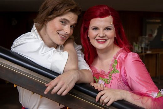 Kaksi värikkäisiin asuihin pukeutunutta ihmistä poseeraa ja hymyilee. Vasemmalla olevalla henkilöllä on pitkät ruskeat hiukset ja valkoinen paita, kun taas oikealla olevalla henkilöllä on kirkkaanpunaiset hiukset ja vaaleanpunainen mekko, jossa on vihreitä korostuksia.