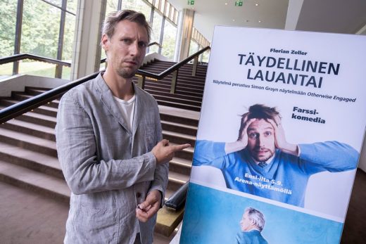 Vaaleanharmaapaitainen mies seisoo sisätiloissa portaiden vieressä ja osoittaa suomalaisen teatterin julistetta, jossa stressaantunut mies pitää päätään. Julisteessa lukee Täydellinen lauantai ja siinä kerrotaan tapahtuman yksityiskohdat.