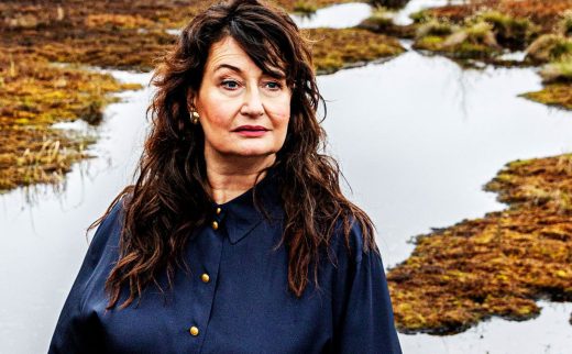 Nainen, jolla on pitkät ruskeat hiukset ja tumma napitettava paita, seisoo ulkona veden ja ruohokenttien läheisyydessä ja katsoo hieman sivulle neutraali ilme kasvoillaan. Taustalla on sammaloitunutta maastoa ja matalia altaita.