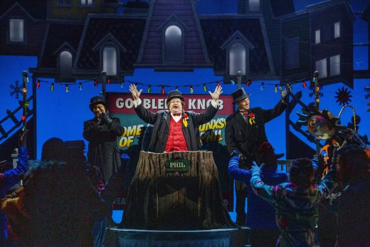 Punaliivinen ja silinterihattuinen mies seisoo kädet ylhäällä korokkeen takana, jossa lukee PHIL, ja häntä reunustaa kaksi tummiin takkeihin ja hattuihin pukeutunutta miestä, kun väkijoukko hurraa-huutoja juhlallisessa, koristellussa ulkoilmatilassa.