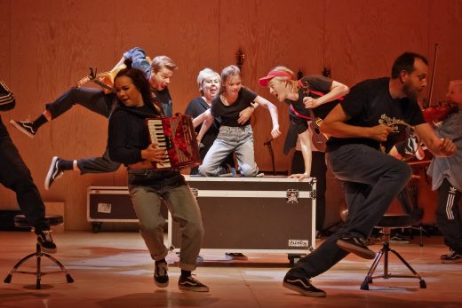 Vilkas esiintyjäjoukko tanssii ja soittaa energisesti soittimia, kuten harmonikkaa ja viulua, lavalla, jossa on puinen tausta. Jotkut hyppäävät ilmaan, kun taas toiset osallistuvat vilkkaasti suuren laitekotelon ympärille.