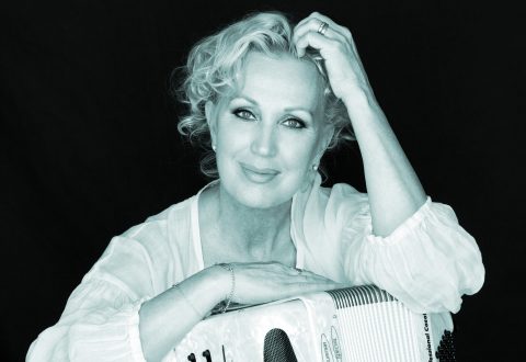 Hymyilevä nainen, jolla on lyhyet, kiharat vaaleat hiukset, nojaa toisella kädellään harmonikkaan ja koskettaa toisella kädellään päätään. Hänellä on vaalea pusero tummalla taustalla.