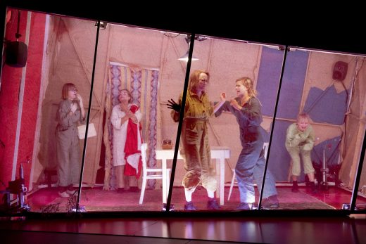 Viisi värikkäisiin haalareihin pukeutunutta ihmistä esiintyy vinossa lavalla, joka on erotettu katsojasta läpinäkyvällä esteellä. He näyttävät näyttelevän intensiivisessä kohtauksessa, joka sijoittuu teltan kaltaiseen huoneeseen, jossa on harvat huonekalut.