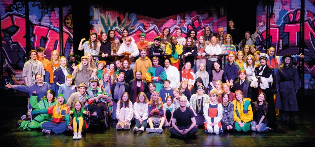 Suuri joukko värikkäisiin asuihin pukeutuneita ihmisiä poseeraa lavalla eloisan, graffititaustan edessä hymyillen.