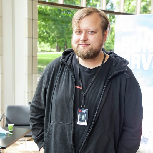 Vaaleanruskeat hiukset ja partainen mies, jolla on musta huppari ja virkamerkki kaulassaan, seisoo sisätiloissa ikkunan ääressä, jonka ulkopuolella näkyy vihreitä puita.