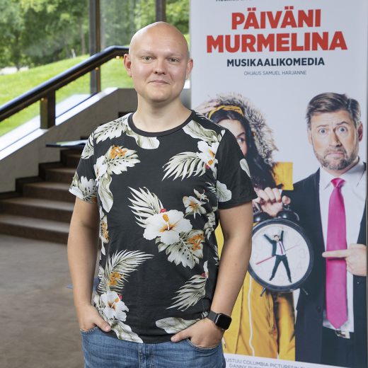 Mustaan kukkapaitaan pukeutunut mies seisoo sisätiloissa ja hymyilee kameralle. Hänen takanaan on Päiväni Murmelina -musikaalikomedian juliste, jossa on kaksi hahmoa, joista toisella on kädessään suuri kello.
