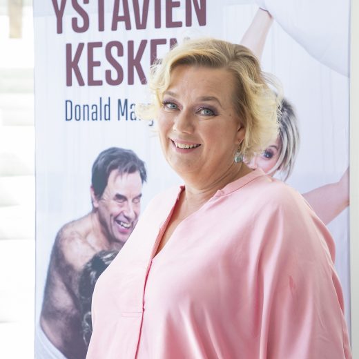 Hymyilevä nainen, jolla on lyhyet vaaleat hiukset ja vaaleanpunainen pusero, seisoo julisteen edessä, jossa on suomenkielistä tekstiä ja taustalla näkyvät osittain kahden ihmisen kasvot.