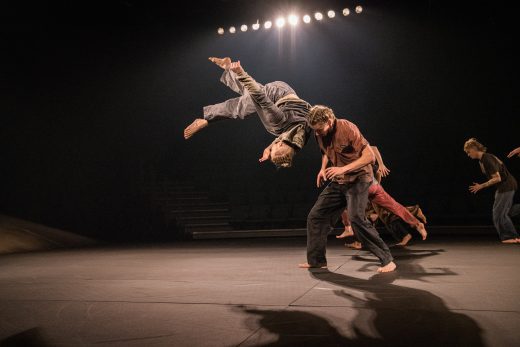 Paljain jaloin tanssiva tanssija on keskellä ilmaa, ja hän kääntyy toisen tanssijan selän yli hämärästi valaistulla näyttämöllä. Taustalla liikkuu useita muita tanssijoita kirkkaiden ylävalojen alla.