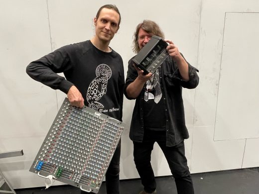 Kaksi ihmistä seisoo sisätiloissa hymyillen ja äänentoistolaitteita kädessään. Toisella on kädessään suuri miksauspöytä ja toisella pienempi elektroninen laite. Tausta on yksinkertainen, valkoinen ja teollisen näköinen.