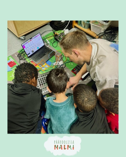 Mies auttaa neljää lasta käyttämään lattialla äänimikseriä ja kannettavaa tietokonetta ja opettaa heille musiikin tuottamista. Lapset katsovat tarkasti, ja heidän ympärillään näkyy värikkäitä kaapeleita ja laitteita.