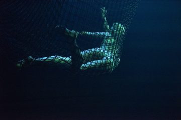 Henkilö on kietoutunut suureen verkkoon, jota valaisee sinivihreä valo pimeässä ympäristössä, mikä luo dramaattisen ja surrealistisen vedenalaisen tunnelman.