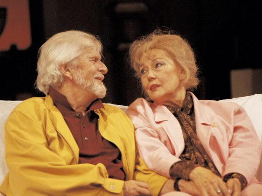 Iäkäs mies ja nainen istuvat lähekkäin sohvalla ja katsovat toisiaan lempein ilmein. Miehellä on keltainen takki ja naisella vaaleanpunainen puku.