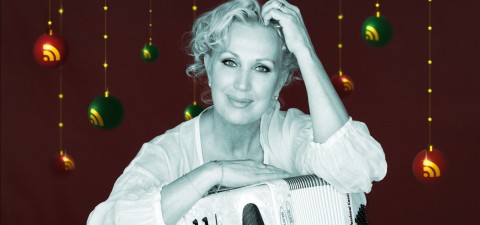 Nainen, jolla on lyhyet, kiharat vaaleat hiukset, hymyilee ja lepuuttaa kättään harmonikan päällä. Hänellä on vaalea pusero, ja taustalla roikkuu juhlavia punaisia ja vihreitä koristeita tummaa taustaa vasten.