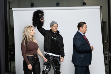 Nainen ruskeissa vaatteissa, mies gorillanaamari käsinukkena ja mies mustassa puvussa seisovat valkoisen kuvausseinän edessä.
