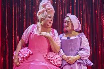 Lavalla seisoo kaksi esiintyjää taidokkaissa puvuissa. Toisella on kirkkaan vaaleanpunainen mekko ja pitkä puuteroitu peruukki, kun taas toisella on lila mekko ja hattu. Molemmilla on dramaattinen meikki, ja taustalla on hohtava punainen verho.