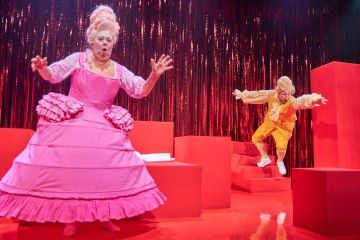 Kaksi näyttelijää esiintyy kirkkaanpunaisella näyttämöllä; toisella on suuri vaaleanpunainen mekko ja valkoinen peruukki, toisella keltainen puku ja peruukki, ja he näyttävät hyppivän tai putoavan dramaattisesti. Taustalla on punaiset verhot ja geometriset lavasteet.