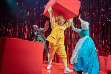 Lavalla esiintyy kolme näyttelijää, joilla on värikkäät puvut; yksi keltapukuinen nostaa suurta punaista laatikkoa yläpuolelle, kun taas toinen sinipukuinen näyttelijä kurottautuu heitä kohti. Kolmas harmaapukuinen näyttelijä ilmestyy säikähtäen taustalle kiiltävää punaista verhoa vasten.