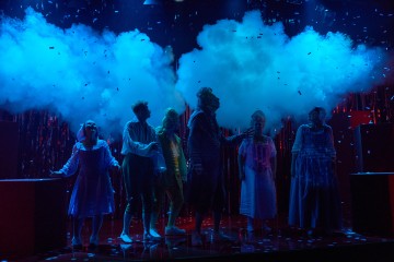 Kuusi teatteripukuista ihmistä seisoo lavalla dramaattisen sinisen valaistuksen alla, ja heidän yläpuolellaan on savu- ja konfettipilviä. Taustalla on pimeää, jossa kimmeltää kimaltava kimalle, mikä luo maagisen, surrealistisen ilmapiirin.