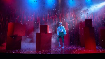 Valkoiseen paitaan ja tummiin housuihin pukeutunut paljasjalkainen henkilö seisoo lavalla, jossa on punaisia kuutioita, ripoteltuja konfetteja ja dramaattinen sininen ja vaaleanpunainen valaistus, jota ympäröi sumu ja pystysuorista viivoista koostuva tausta.