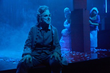 Historialliseen pukuun pukeutunut vanhempi henkilö istuu sinisen näyttämövalaistuksen alla mietteliään näköisenä. Taustalla seisoo kolme muuta aikakauden vaatteisiin ja hattuihin pukeutunutta näyttelijää, jotka ovat osittain piilossa pylvään takana, ja lattialle on ripoteltu konfetteja.