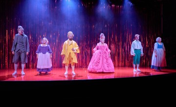 Kuusi näyttelijää värikkäissä, liioitelluissa aikakauden puvuissa seisoo paljain jaloin kirkkaanpunaisella näyttämöllä dramaattisessa sinisessä valaistuksessa, jonka taustalla on hohtavan punainen verho.