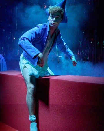 Siniseen terävään hattuun ja takkiin pukeutunut henkilö istuu punaisella korokkeella toinen jalka kohotettuna sinisen valaistuksen ja teatterisavun ympäröimänä, mikä luo dramaattisen ja surrealistisen tunnelman.