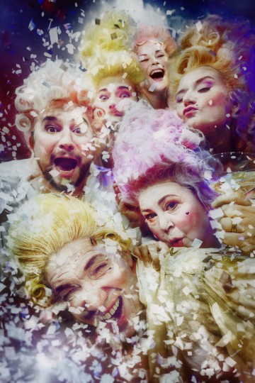 Kuusi ihmistä, joilla on taidokkaat, värikkäät 1700-luvun peruukit ja puvut, nauravat ja juhlivat pudottavan konfetin ympäröimänä eloisaa, juhlavaa taustaa vasten.