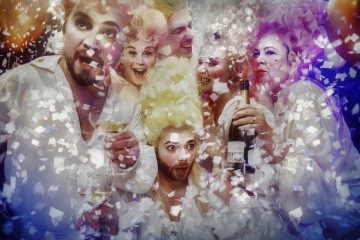 Ryhmä ihmisiä, jotka ovat pukeutuneet ylellisiin 1700-luvun tyyliin sopiviin pukuihin, juhlii konfettien, viinin ja samppanjan kanssa. Heidän kasvoillaan näkyy yllätystä ja innostusta juhlatunnelman keskellä.