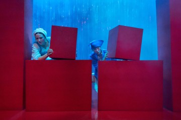 Kaksi esiintyjää, toinen valkoisissa ja toinen sinisissä puvuissa, kyyristelevät lavalla suurten punaisten kuutioiden takana, ja sininen, hohtava tausta viittaa teatteri- tai draamaympäristöön.