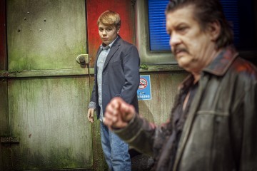 Blazeriin ja farkkuihin pukeutunut nuori mies seisoo huolestuneen näköisenä vanhentuneen, vihreän ja punaisen seinän vieressä. Etualalla näkyy epäselvästi nahkatakkinen vanhempi mies, jonka nyrkki on osittain koholla.