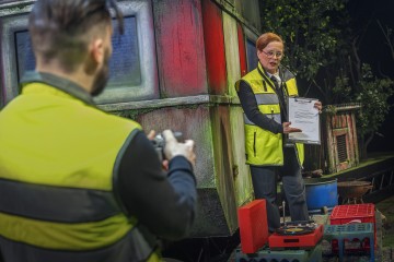 Keltaisiin turvaliiveihin pukeutunut nainen pitää kädessään asiakirjaa ja puhuu miehelle, jolla on myös turvaliivit ja jolla on kamera kädessään. He ovat ulkona lähellä säästä kärsivää rakennusta, ja taustalla on erilaisia laitteita ja puita.