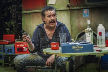 Viiksekäs mies istuu sotkuisen pöydän ääressä pelikortit kädessään. Pöydällä on pulloja, muki ja hajallaan papereita. Hän näyttää puhuvan tai reagoivan johonkin ilmeikkäästi. Ympäristö vaikuttaa maalaismaiselta ja kuluneelta.