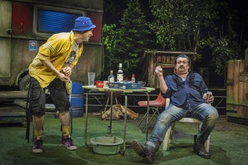 Kaksi miestä on vuorovaikutuksessa ulkona pöydän ääressä, jossa on juomia ja välipaloja. Toinen, keltainen paita ja sininen hattu, nojaa eteenpäin. Toinen, sinipaitainen, istuu savuke kädessään ja näyttää yllättyneeltä. Taustalla on asuntovaunu ja puita.