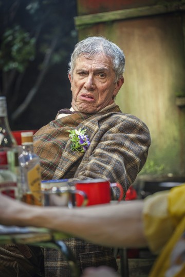 Iäkäs ruudulliseen takkiin pukeutunut mies, jolla on kukka kauluksessa, tekee inhottavan ilmeen istuessaan ulkona pöydän ääressä, jossa on pulloja ja mukeja, ja taustalla on viherkasveja ja puurakennelma.