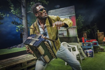 Mies, jolla on aurinkolasit ja ruskea takki, soittaa harmonikkaa energisesti ulkolavalla, jossa on muovituoleja, puulankkuja, puita ja pieni rakennus taustalla.