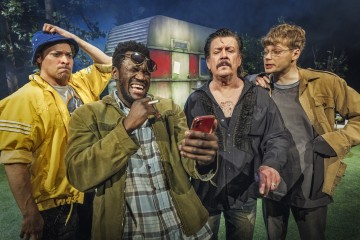 Neljä miestä seisoo ulkona ränsistyneen perävaunun edessä. Yksi mies nauraa ja osoittaa puhelinta, kun muut katselevat vaihtelevin uteliaisuuden ja huvittuneisuuden ilmein. He ovat pukeutuneet rentoihin, karkeisiin vaatteisiin.