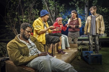 Viisi värikkäisiin, rentoihin vaatteisiin pukeutunutta ihmistä on kokoontunut metsämaisemaan. Yksi henkilö istuu penkillä, kun taas muut seisovat tai istuvat, keskustelevat vilkkaasti ja pitävät kädessään kortteja tai papereita. Heidän ympärillään on puita ja kasveja.