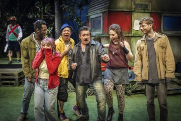 Kuusi ihmistä seisoo yhdessä ulkona ilmeikkäissä asennoissa ja vaikuttaa innostuneilta tai yllättyneiltä. Heidän takanaan on värikkääseen asuun pukeutunut henkilö ja rakennus, jossa on punaisia ja vihreitä paneeleita ja jota ympäröi vehreys ja puulankut.