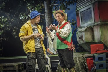 Kaksi värikkäisiin pukuihin pukeutunutta näyttelijää, joista toisella on sininen hattu ja keltainen takki ja toisella kansanomainen asu, jossa on olkihattu ja punainen vyö, seisovat vilkkaasti keskustelemassa puiden ja maalaismaiseman vaunun muodostaman lavastuksen edessä.