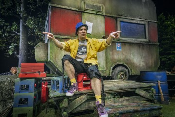 Siniseen hattuun ja keltaiseen takkiin pukeutunut henkilö istuu puuportailla ränsistyneen asuntovaunun edessä ja osoittaa molemmilla käsillään ulospäin. Lavastuksessa on laatikoita, työkaluja ja vihreitä lehtiä, mikä antaa maalaismaisen ulkoilman tunnelman.