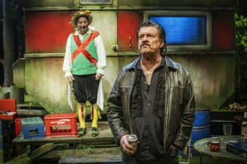 Mustaan nahkatakkiin pukeutunut mies pitelee tölkkiä ja seisoo värikkään, eksentrisen, liiviin, hattuun ja kirkkaisiin sukkiin pukeutuneen hahmon edessä, jonka taustalla on perävaunu ja erilaisia esineitä.