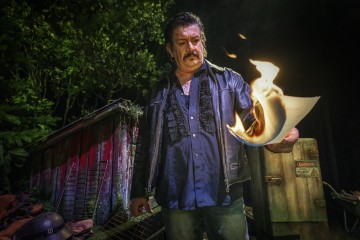 Siniseen paitaan ja nahkatakkiin pukeutunut mies pitää kädessään palavaa paperiarkkia, kun hän seisoo ulkona yöllä lähellä ränsistynyttä hökkeliä ja umpeenkasvanutta lehtimetsää. Hänen ilmeensä on vakava ja intensiivinen.