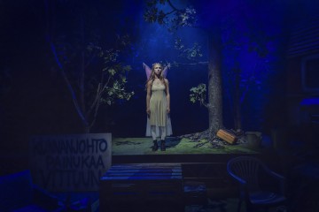 Vaaleaan mekkoon ja keijusiipiin pukeutunut nainen seisoo sinisten näyttämövalojen alla metsäisessä lavasteessa. Hänen ympärillään on puita, lehtiä ja hajallaan olevaa rekvisiittaa. Etualalla näkyy suomenkielinen kyltti.