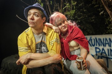 Keltaiseen takkiin ja siniseen hattuun pukeutunut mies istuu vaaleanpunaiset hiukset ja punainen huivi kantavan naisen vieressä, joka nojaa miehen olkapäähän tölkki kädessään. He vaikuttavat mietteliäiltä, taustalla näkyy puita ja graffiteja.