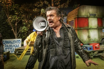 Mustaan nahkatakkiin pukeutunut mies huutaa megafoniin ulkoilmaesityksen aikana, taustalla kyltti, puita ja asuntoauto. Hän vaikuttaa eloisalta ja intensiiviseltä.